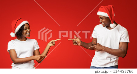 Emotional african american young man and woman in Santa hats pointing at copy space on orange studio background, showing advertisement for New Year or Christmas deal, offer, sale, discount Emotional african american young man and woman in Santa hats pointing at copy space on orange studio background, showing advertisement for New Year or Christmas deal, offer, sale, discount 134455766