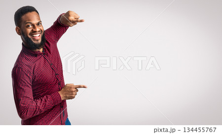 Smiling African Guy Pointing Fingers Aside Showing Blank Space Standing Over White Background, Studio Shot. Look There, Great Offer Advertisement. Black Man Advertising Your Text 134455767