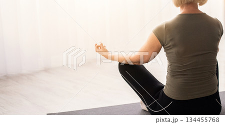 Back view of peaceful senior woman meditating in lotus pose at home, empty space. Calm mature lady doing morning yoga practice, facing window, making gyan mudra, reaching zen 134455768