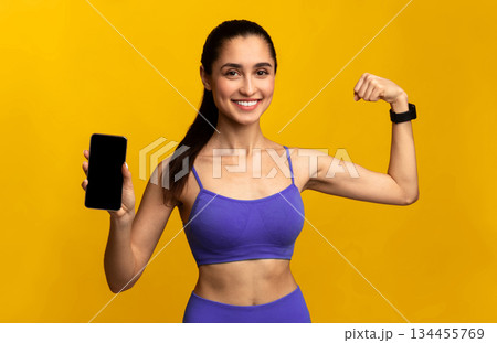 Sports App And Healthy Lifestyle. Positive slim lady wearing blue sportswear holding smartphone with black blank screen for mockup, showing her biceps, isolated on orange background, studio shot 134455769