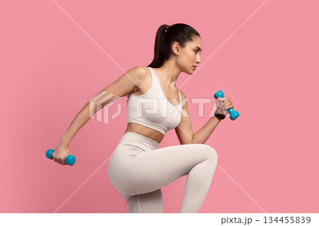 Training Concept. Full Length Portrait Of Confident Fit Young Lady In Sportswear Exercising With Two Dumbbells And Lifting Leg Up, Isolated On Pink Studio Background. Energy And Sports Training Concept. Full Length Portrait Of Confident Fit Young Lady In Sportswear Exercising With Two Dumbbells And Lifting Leg Up, Isolated On Pink Studio Background. Energy And Sports 134455839