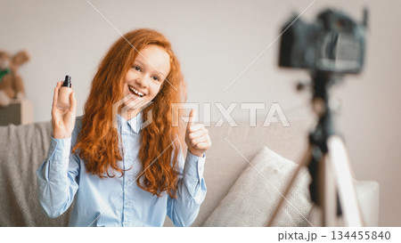 Beauty Advice And Blog. Smiling female teen doing makeup, recording cosmetics product review, showing her foundation for young skin to camera on tripod. Girl filming video, showing thumbs up 134455840