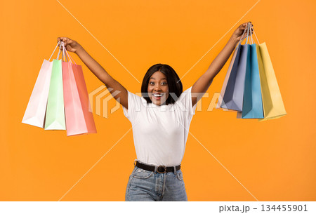 Emotional african american young woman raising hands up with colorful shopping bags and screaming cheerfully, celebrating sales season beginning over orange studio background 134455901