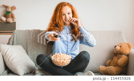 People And Leisure Concept. Portrait of smiling young teenage girl watching tv, eating popcorn from glass bowl in her living room, holding remote control, resting and relaxing sitting on the couch 134455926
