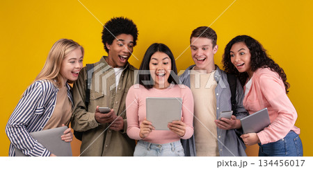 Modern gadgets, social networks and education. Shocked cheerful young multi ethnic students looking in digital tablet in asian female hands, with open mouths, isolated on orange background, copy space 134456017