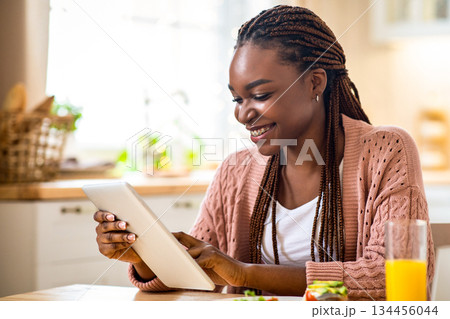 Young Smiling Black Woman Using Digital Tablet During Breakfast In Kitchen. Happy African American Lady Eating Tasty Food And Browsing Internet, Shopping Online Or Checking Social Networks, Free Space 134456044