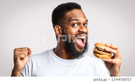 Joyful Black Guy Biting Burger Enjoying Cheat Meal Eating Unhealthy Cheeseburger Standing Posing Over White Studio Background. Junk Food Overeating, Obesity Problem And Unhealthy Nutrition Habit 134456057