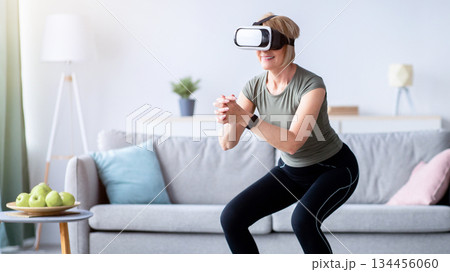 Exercising with virtual reality concept. Athletic mature woman in VR headset doing squats on her home workout. Positive senior lady using modern technologies for domestic training Exercising with virtual reality concept. Athletic mature woman in VR headset doing squats on her home workout. Positive senior lady using modern technologies for domestic training 134456060