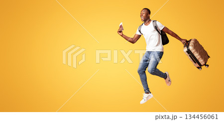 Finally vacation. Young happy black man with suitcase, backpack, passport and flight tickets jumping up orange studio background. Excited african american guy running towards copy space, panorama 134456061