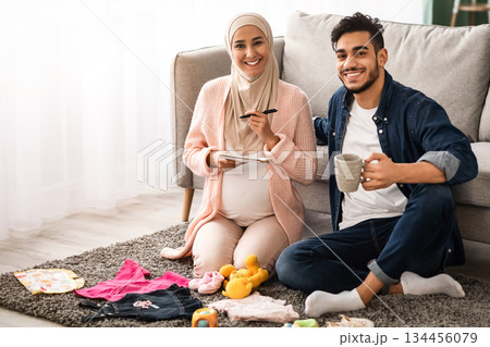 Portrait Of Happy Muslim Couple Making Preparations While Awaiting A Baby, Cheerful Pregnant Middle Eastern Spouses Sitting Next To Child Clothes At Home, Preparing Checklist For Hospital, Free Space Portrait Of Happy Muslim Couple Making Preparations While Awaiting A Baby, Cheerful Pregnant Middle Eastern Spouses Sitting Next To Child Clothes At Home, Preparing Checklist For Hospital, Free Space 134456079