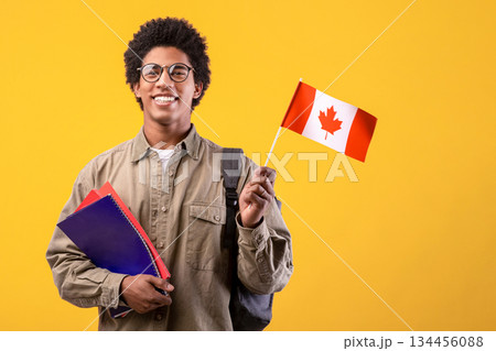 Education, school, college and university. Happy young african american guy in glasses holding notepads and small Canada flag and ready to study, isolated on orange background, free space, studio shot 134456088