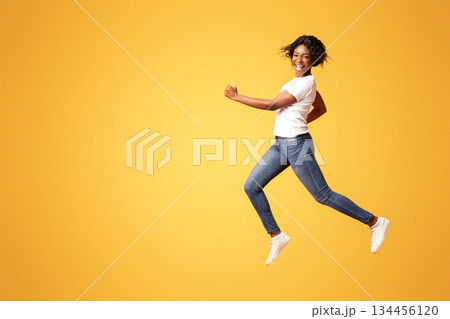 Active black lady in casual outfit running over orange studio background, full length shot. Cheerful african american young woman in jeans and white t-shirt having fun on her own 134456120