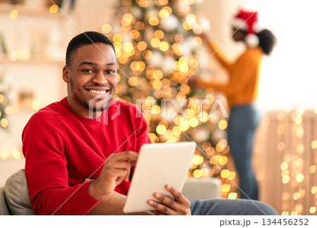 Christmas eve shopping online during sale and discount, and video call to family. Smiling millennial african american man typing on tablet, woman decorating tree in living room interior to garlands Christmas eve shopping online during sale and discount, and video call to family. Smiling millennial african american man typing on tablet, woman decorating tree in living room interior to garlands 134456252