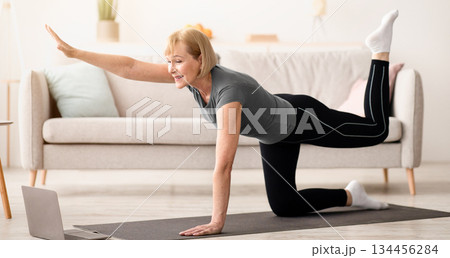 Home sports. Fit senior woman working out in front of laptop, lifting her leg and arm in living room. Athletic mature lady exercising on yoga mat, doing domestic fitness to online training program 134456284