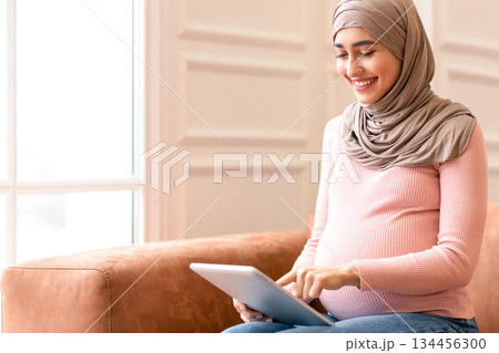 Happy Pregnant Muslim Woman Using Digital Tablet Reading Blog About Pregnancy Online Sitting On Sofa At Home. Middle Eastern Female In Hijab Browsing Internet. People And Technology Concept Happy Pregnant Muslim Woman Using Digital Tablet Reading Blog About Pregnancy Online Sitting On Sofa At Home. Middle Eastern Female In Hijab Browsing Internet. People And Technology Concept 134456300