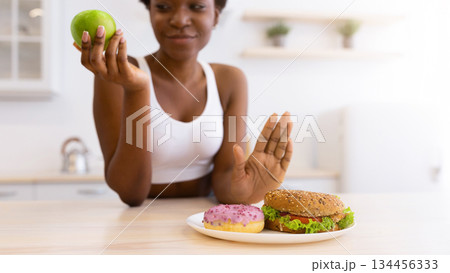 Black Woman Refusing To Eat Unhealthy Burger And Donut Choosing Healthy Apple Fruit Sitting In Modern Kitchen At Home. No Cheat Meal, Stop Eating Junk Food Concept. Selective Focus, Shallow Depth 134456333