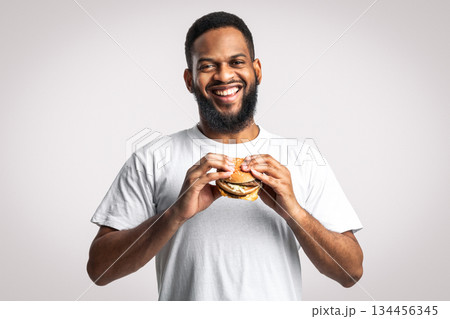 Junk Food. Happy Black Man Holding Burger Standing On White Background In Studio, Smiling Looking At Camera. Nutrition, Overeating Habit And Unhealthy Eating Concept Junk Food. Happy Black Man Holding Burger Standing On White Background In Studio, Smiling Looking At Camera. Nutrition, Overeating Habit And Unhealthy Eating Concept 134456345