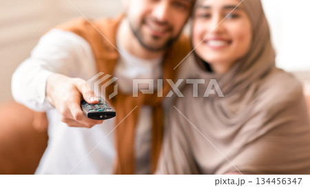Cheerful Muslim Couple Watching Movie On TV Sitting Together On Sofa At Home, Focus On Television Remote Control. Arab Husband And Wife Switching Channels Looking At Camera. Shallow Depth Cheerful Muslim Couple Watching Movie On TV Sitting Together On Sofa At Home, Focus On Television Remote Control. Arab Husband And Wife Switching Channels Looking At Camera. Shallow Depth 134456347