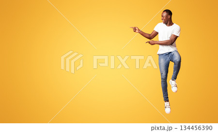 Excited black guy jumping up and showing copy space over orange studio background. Emotional african american millennial handsome man smiling and pointing at advertising or announcement 134456390