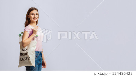 Smiling Young Woman Carrying Eco Tote Bag With No More Plastic Inscription Filled With Empty Bottles Ready For Recycling, Millennial Lady Walking Over Light Studio Background, Copy Space 134456425