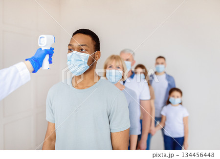 Coronavirus immunization medical campaign concept. Black man in face mask standing in line at clinic, doctor measuring his temperature with infrared thermometer before covid-19 vaccination, copy space 134456448