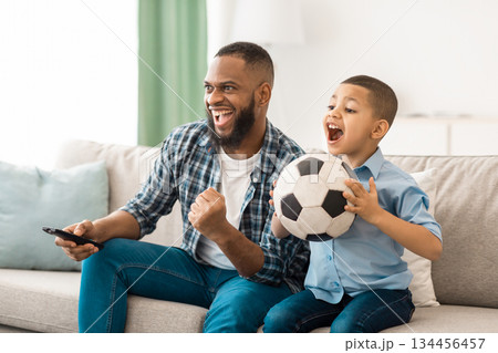 Happy African American Father And Son Watching Sports On TV Holding Soccer Ball And Remote Control Sitting On Sofa At Home. Family Watches Television. Sport TV Programming Concept 134456457