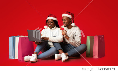 Christmas shopping. Cheerful black couple buying gifts online, using laptop and credit card, happy african spouses in santa hats sitting on orange background with lots of bright shopper bags, panorama 134456474