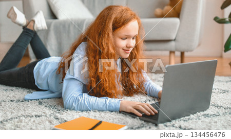 Browsing Internet. Portrait of red-haired girl lying on the floor carpet in living room, using laptop computer, watching video or movie, chatting with friends, surfing web and smiling Browsing Internet. Portrait of red-haired girl lying on the floor carpet in living room, using laptop computer, watching video or movie, chatting with friends, surfing web and smiling 134456476