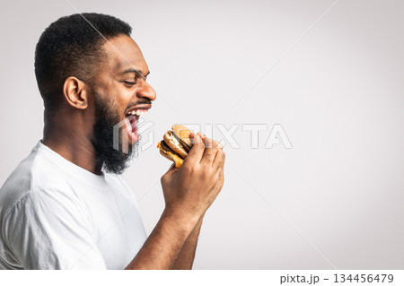 Side View Of Hungry Black Man Eating Burger Posing Standing Over White Background, Studio Shot. Free Space For Your Text. Junk Food, Unhealthy Male Nutrition And Overeating Habit. Side View Of Hungry Black Man Eating Burger Posing Standing Over White Background, Studio Shot. Free Space For Your Text. Junk Food, Unhealthy Male Nutrition And Overeating Habit. 134456479