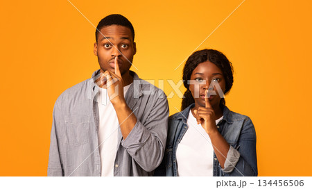 Dont Say A Word. Black man and woman keeping fingers on lips, making silence sign and shh gesture while looking at camera together, young african couple standing isolated on yellow background 134456506