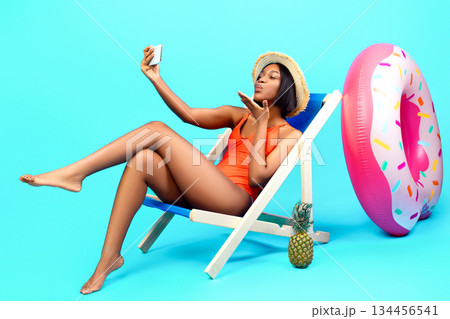 Flirty black woman chatting with her boyfriend on smartphone, blowing air kiss at webcam, making selfie while sitting in lounge chair on beach, blue studio background. Full length portrait 134456541