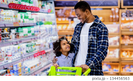Happy black dad and his pretty little daughter with shopping trolley having great time buying groceries at big mall. African American parent with kid purchasing foodstaff at hypermarket 134456563
