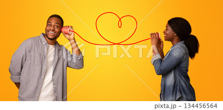 Love Message. Happy black woman whispering romantic words to her boyfriend via tin phone, standing isolated over yellow background, creative collage for Valentine's day with drawn red heart, panorama 134456564