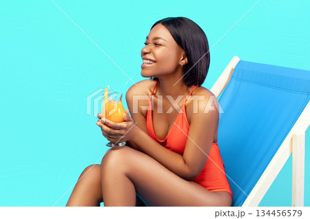 Cheerful young black woman relaxing in lounge chair with delicious fruit cocktail, laughing and enjoying her tropical summer vacation on blue studio background, banner design with copy space Cheerful young black woman relaxing in lounge chair with delicious fruit cocktail, laughing and enjoying her tropical summer vacation on blue studio background, banner design with copy space 134456579