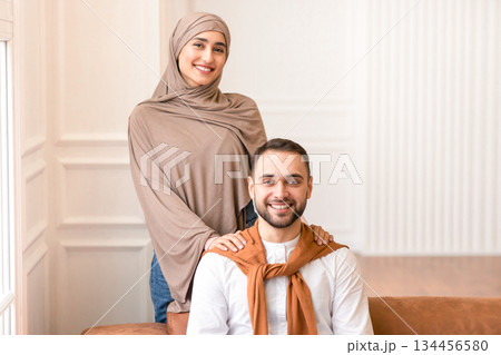 Married Middle Eastern Couple Posing Together Smiling To Camera At Home. Gladful Muslim Wife Wearing Hijab Hugging Her Husband Indoors. Modern Arabic Family Portrait Concept Married Middle Eastern Couple Posing Together Smiling To Camera At Home. Gladful Muslim Wife Wearing Hijab Hugging Her Husband Indoors. Modern Arabic Family Portrait Concept 134456580