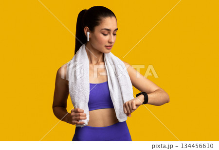 Workout And Electronics Concept. Portrait of fit young sportswoman wearing bracelet, checking fitness tracker on her wrist, listening to music in wireless earphones standing over orange background Workout And Electronics Concept. Portrait of fit young sportswoman wearing bracelet, checking fitness tracker on her wrist, listening to music in wireless earphones standing over orange background 134456610