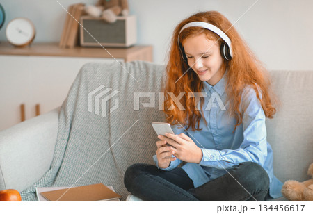 Leisure Concept. Smiling Red-Haired Girl Sitting On The Sofa At Home, Listening To Music In Wireless Headphones, Watching Movie Or Video, Chatting On Her Smartphone, Taking Break From Study Leisure Concept. Smiling Red-Haired Girl Sitting On The Sofa At Home, Listening To Music In Wireless Headphones, Watching Movie Or Video, Chatting On Her Smartphone, Taking Break From Study 134456617