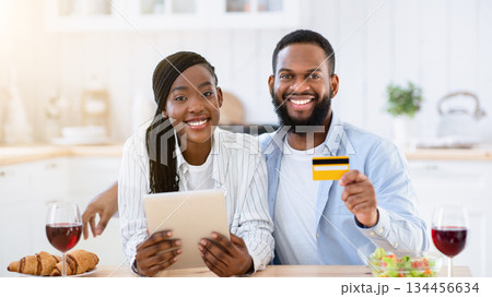 Portraif Of Happy Black Couple With Digital Tablet And Credit Card In Kitchen, Young African Spouses Making Food Or Groceries Delivery Order, Sitting At Table With Red Wine Glasses, Smiling At Camera 134456634