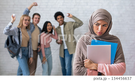Peer problems, victim, racism, humiliation and bullying in college, school or university. Multiracial young students laugh and throw paper at upset arabian woman with books, studio shot, empty space 134456637