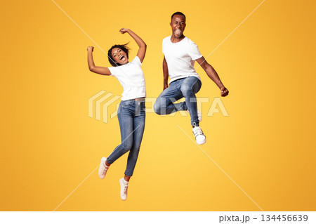 Emotional black millenial couple jumping up on orange studio background, full length shot. Young cheerful african american man and woman celebrating success, having fun together 134456639