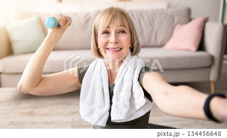 Joyful mature woman taking selfie while exercising with dumbbells at home. Happy senior lady making photo of herself during domestic strength workout, enjoying her training Joyful mature woman taking selfie while exercising with dumbbells at home. Happy senior lady making photo of herself during domestic strength workout, enjoying her training 134456648