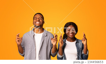 Make A Wish. Superstitious Black Couple Standing With Crossed Fingers And Closed Eyes, African American Man And Woman Pleading For Luck While Standing Together Isolated Over Yellow Background 134456653