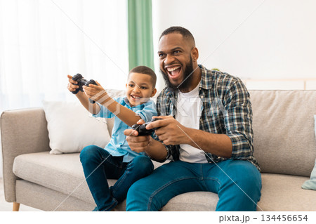 Joyful African Father And His Kid Boy Playing Videogames Bonding And Spending Time Together Having Fun On Weekend At Home. New Video Game Advertisement Concept Joyful African Father And His Kid Boy Playing Videogames Bonding And Spending Time Together Having Fun On Weekend At Home. New Video Game Advertisement Concept 134456654