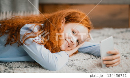 Homeland Generation. Portrait of happy smiling young teenage girl holding and using smartphone while lying on the floor carpet in bedroom, resting and relaxing at home, looking at camera, chatting 134456731