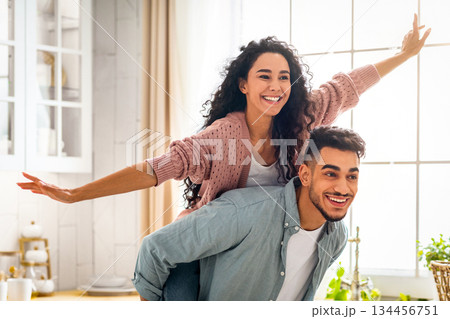 Joyful Middle Eastern Spouses Having Fun Together In Kitchen Interior, Handsome Arab Man Piggybacking His Cheerful Wife, Happy Attractive Lady Enjoying Ride, Spreading Hands Like Flying, Copy Space 134456751