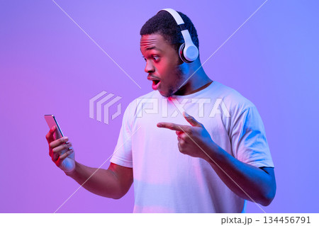 Amazed Black Guy In Wireless Headphones Pointing At Smartphone In His Hands, Excited African American Man Emotionally Reacting To Mobile Offer, Standing In Neon Lighting Over Purple Background Amazed Black Guy In Wireless Headphones Pointing At Smartphone In His Hands, Excited African American Man Emotionally Reacting To Mobile Offer, Standing In Neon Lighting Over Purple Background 134456791