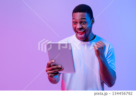 Online Win. Joyful African American Guy Celebrating Success With Digital Tablet, Emotional Happy Black Man Raising Fists And Exclaiming With Excitement, Standing In Neon Light Over Purple Background 134456792