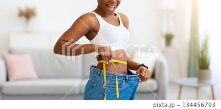 Successful Weight Loss. Joyful Young Black Lady Measuring Waist Wearing Oversized Jeans After Slimming, Smiling To Camera Standing Indoors. Weight-Loss Comparison And Motivation 134456793