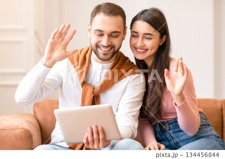 Arab Young Couple Video Calling Using Digital Tablet Computer Waving Hands To Webcam Communicating Online Sitting On Sofa At Home. Modern Remote Communication Concept 134456844