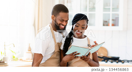 Happy black couple reading recipe in cookbook while cooking healthy vegetarian lunch in kitchen together, african american spouses checking ingredients for tasty vegetable salad, free space Happy black couple reading recipe in cookbook while cooking healthy vegetarian lunch in kitchen together, african american spouses checking ingredients for tasty vegetable salad, free space 134456863